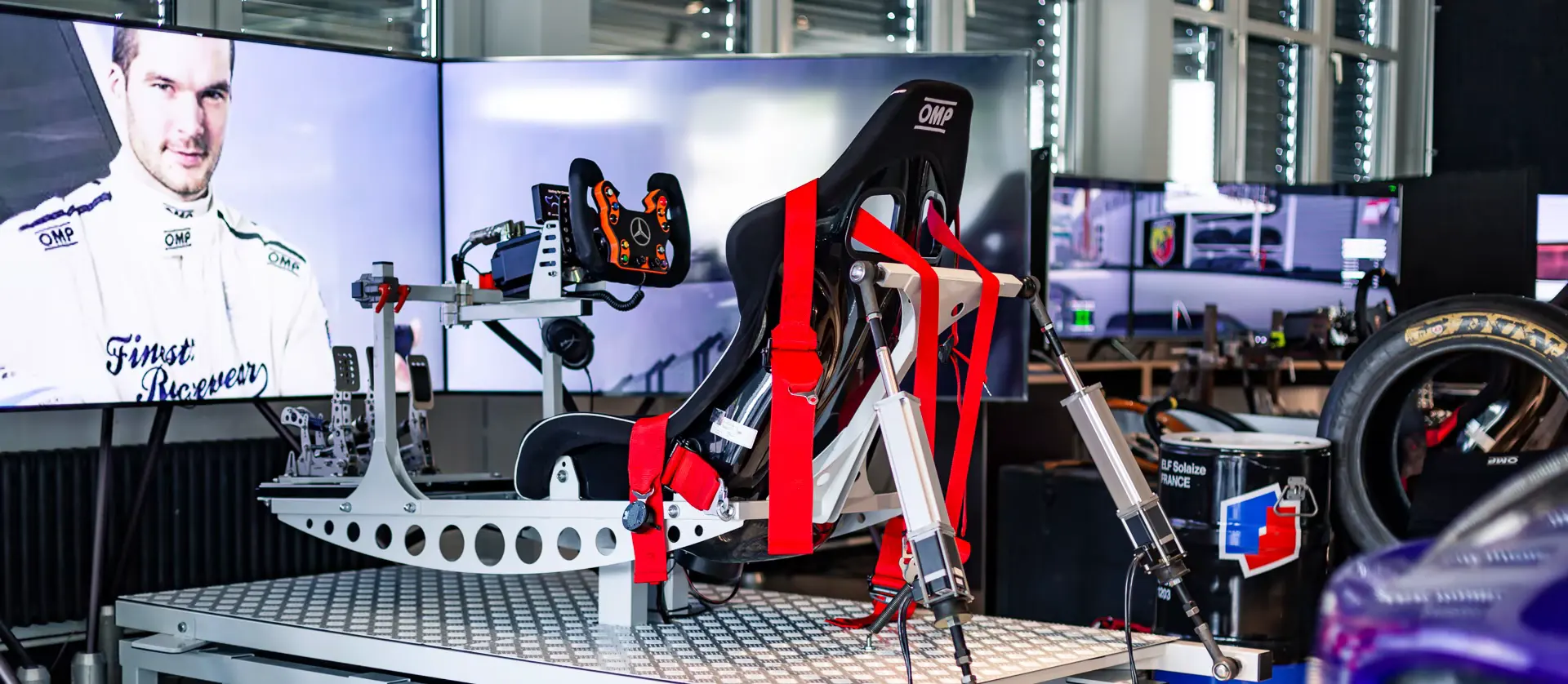 Racing simulator rig with seat, harness and steering wheel in front of its screens